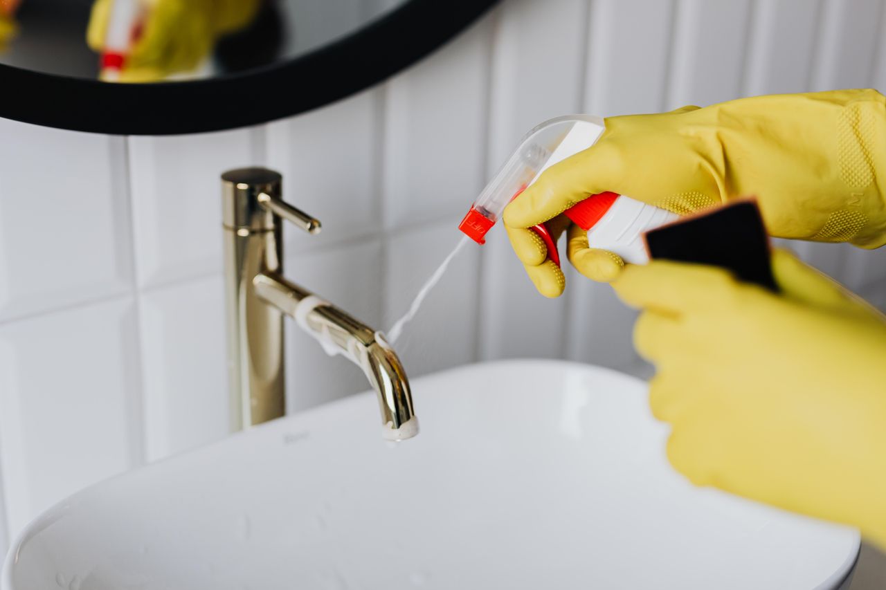 heros-img Seen from above, an anonymous housekeeper in yellow rubber gloves sprays cleaner onto a chrome bathroom water faucet